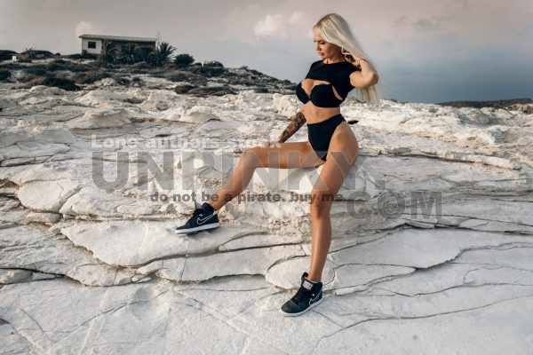 Beautiful sexy woman with waving hairs is sitting on the top of rock near ocean. Lady is resting during sunset at the resort