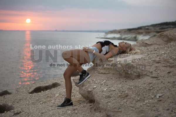 Beautiful sexy woman with waving hairs is sitting on the top of rock near ocean. Lady is resting during sunset at the resort