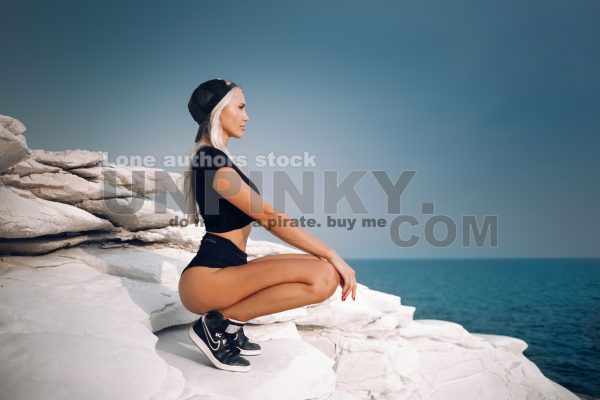 Beautiful woman with waving hairs is sitting on the top of rock near ocean