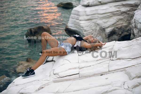 Beautiful sexy woman with waving hairs is sitting on the top of rock near ocean. Lady is resting during sunset at the resort