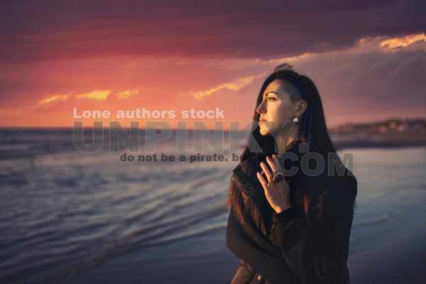 A woman enjoys the sunset at the seaside on a cold day. Portrait of a young lady on the beach during sunset. Lady walks on a cold evening